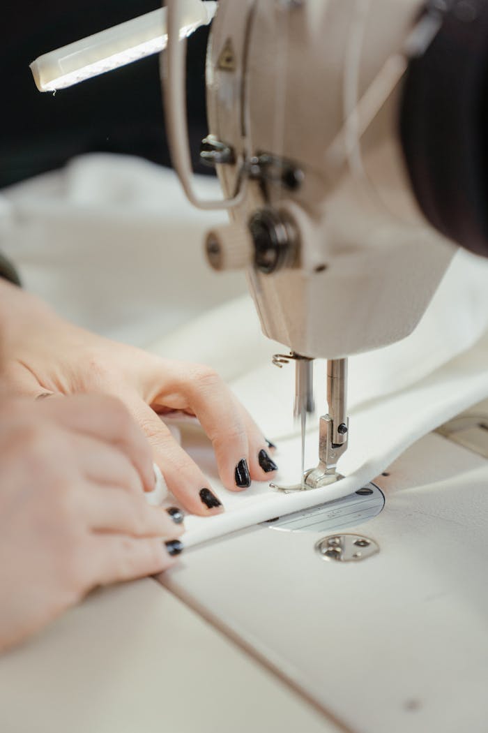 Detailed view of a sewing machine stitching fabric with visible hands, nails painted black.